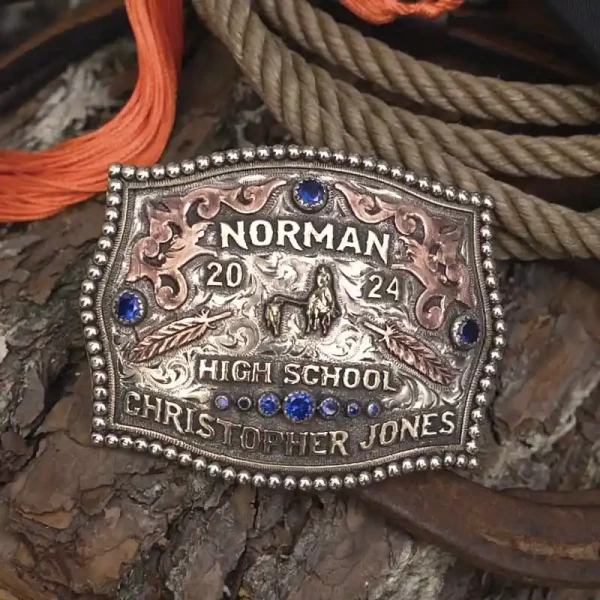 A custom rodeo belt buckle for Norman High School with personalized name featuring feathers and a calf roping figure and blue zirconia stones